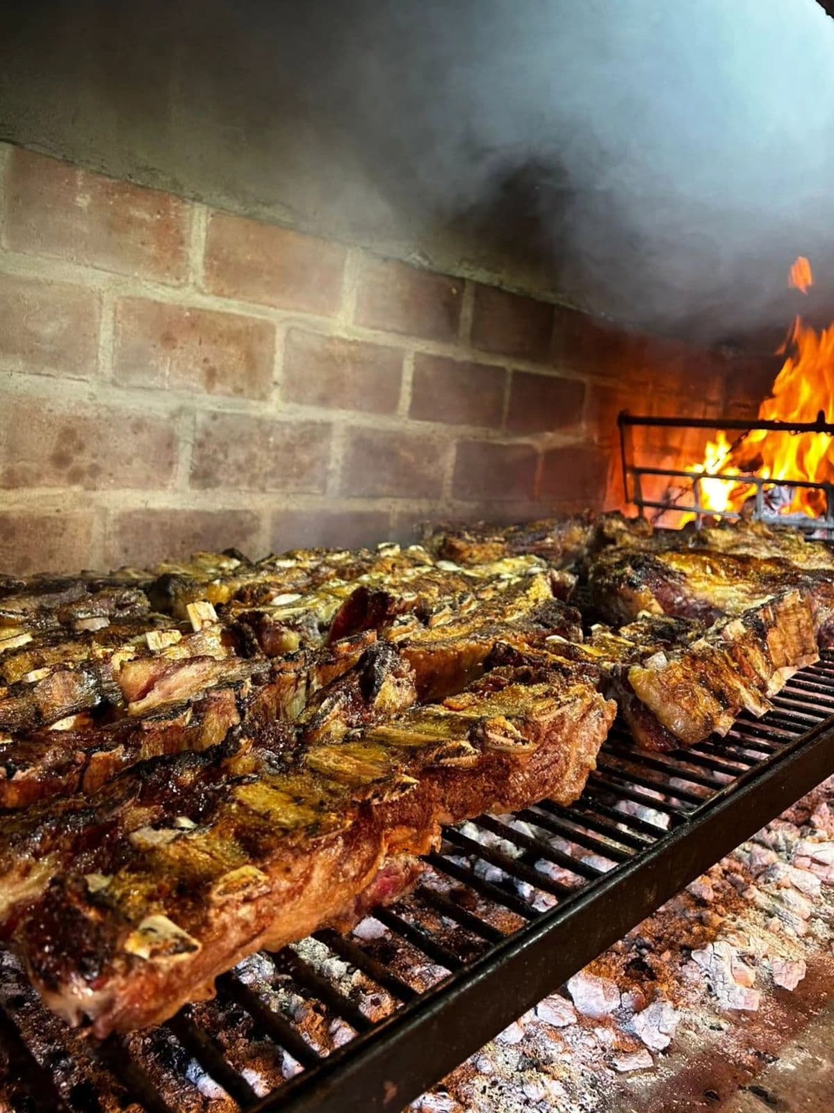 Costillar dorado sobre la parrilla de un quincho de ladrillo, con fuego de leña al fondo