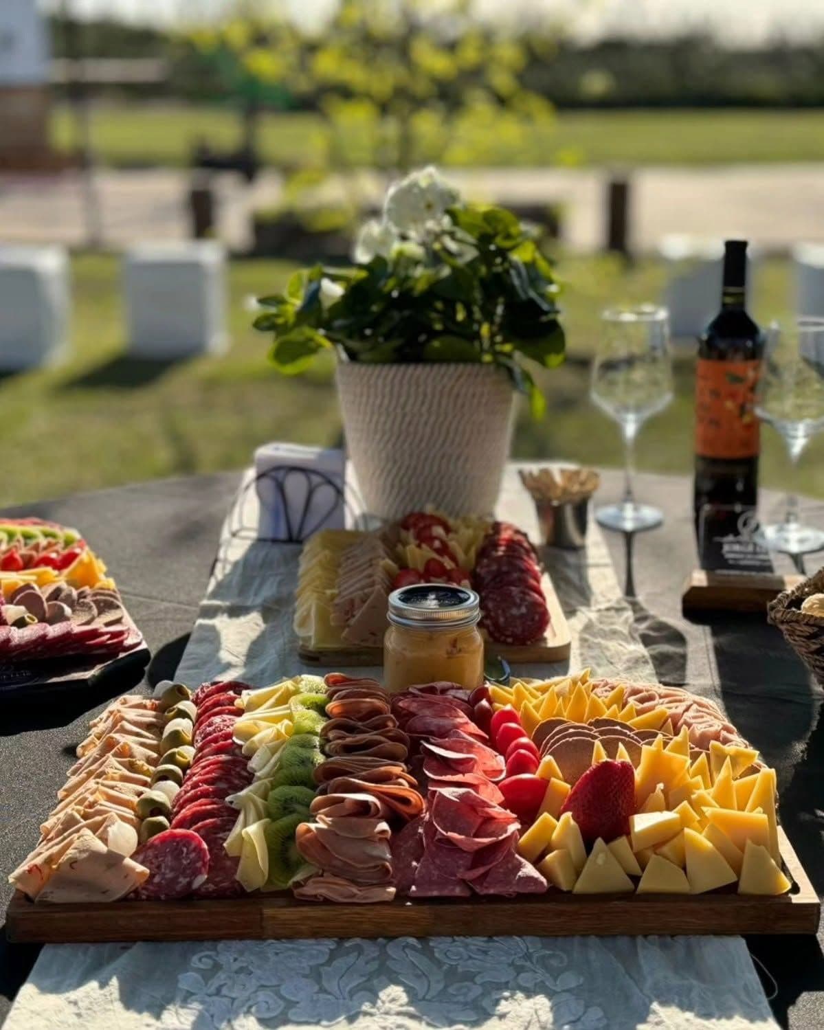 Tabla de fiambres, quesos, kiwi, frutillas y tomates cherry en una mesa al aire libre, con vino y flores