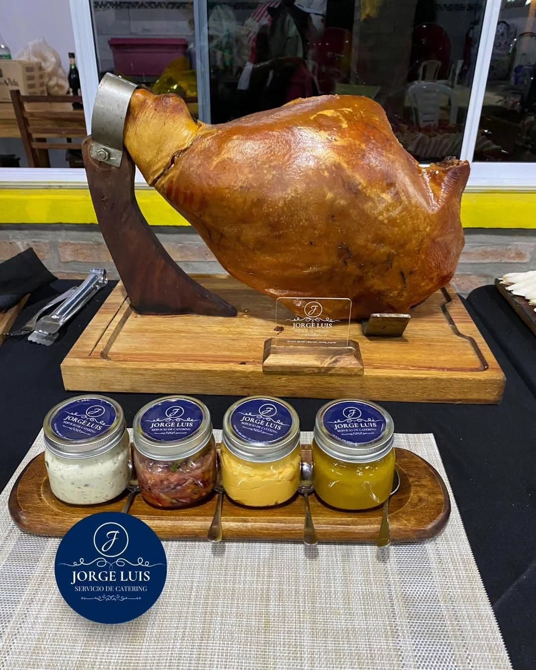Pernil asado entero en un soporte de madera, con cuatro frascos de salsas con la etiqueta de Jorge Luis