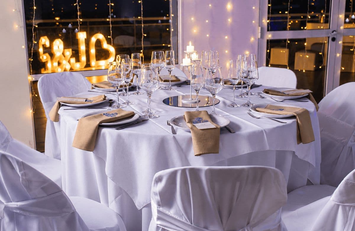 Mesas redondas con manteles blancos, servilletas beige y sillas vestidas de blanco