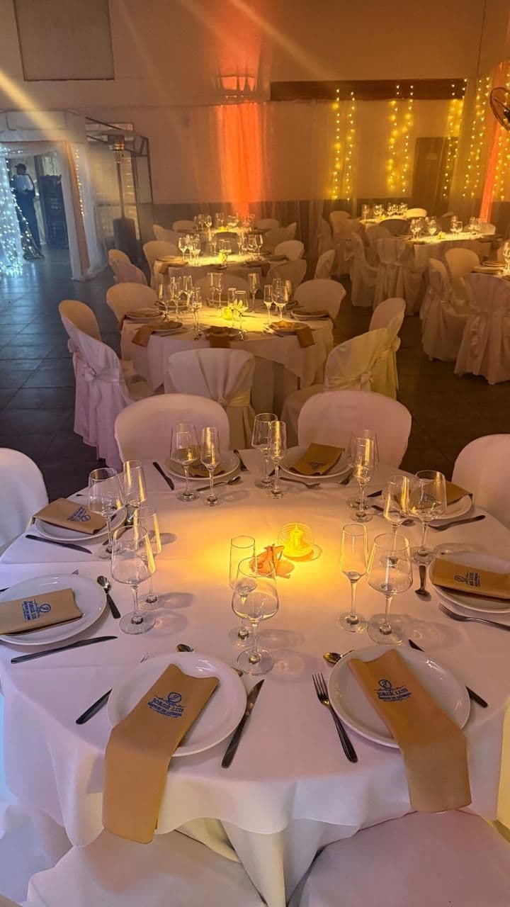 Salón con mesas redondas de mantel blanco, sillas con funda blanca y moño dorado, copas y servilletas de Jorge Luis